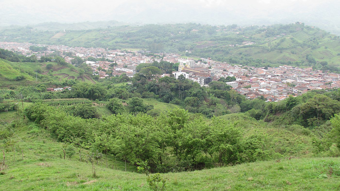 Casco urbano entre colinas de pasto, con una vaguada arbolada en primer plano separando el borde construido del suelo rural